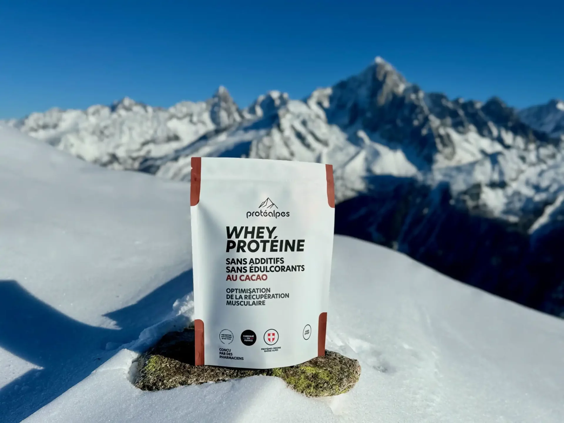 Sac de Whey protéine Protéalpes dans la neige, montagne en arrière-plan. Produit sans additifs ni édulcorants.