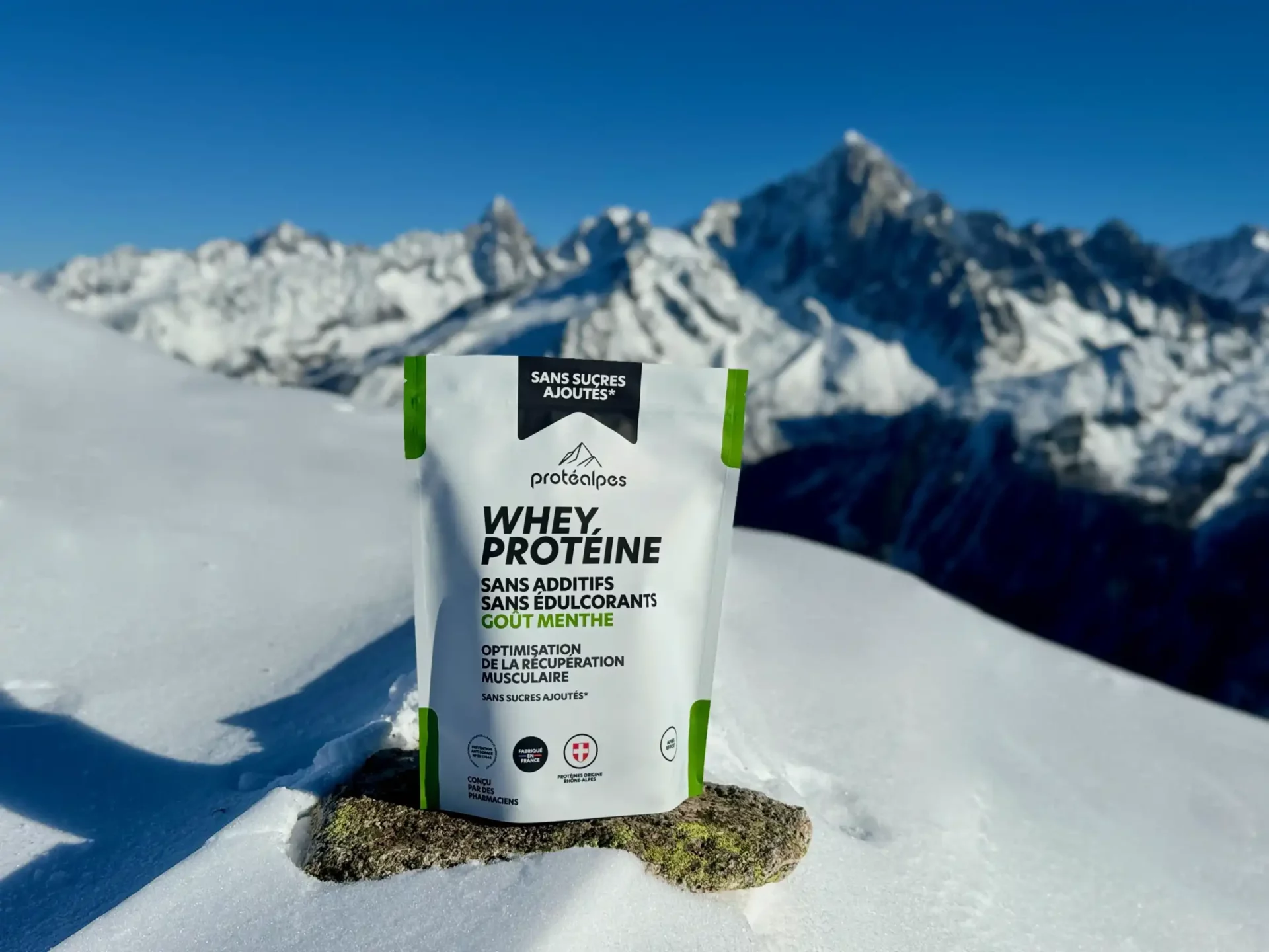Whey protéine menthe Protéalpes sans additifs ni édulcorants, posée sur neige avec montagnes en arrière-plan.
