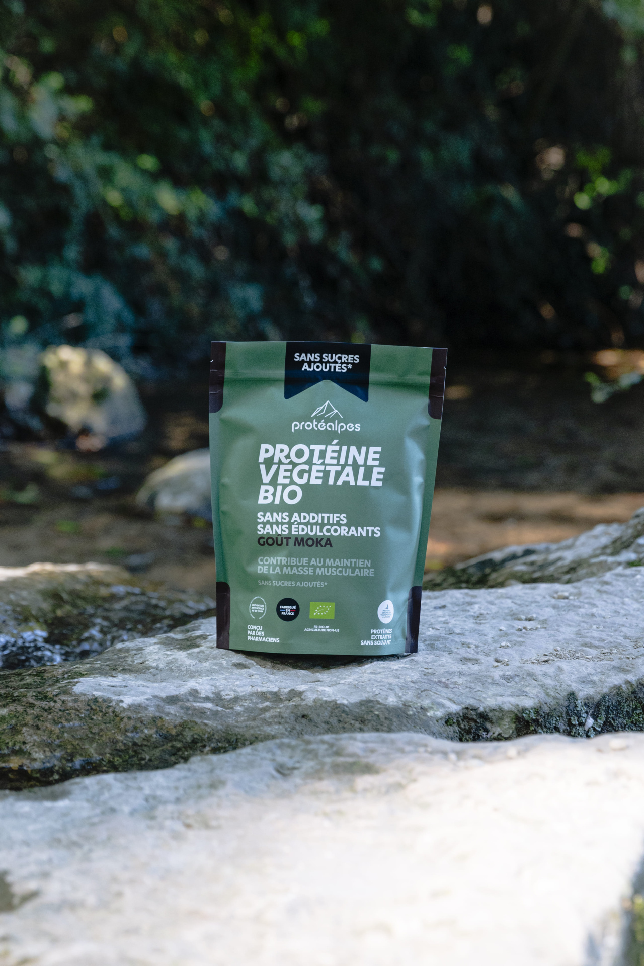Sac de protéine végétale bio de Protéalpes, sans additifs ni édulcorants, goût moka, sur un rocher près d'une rivière.